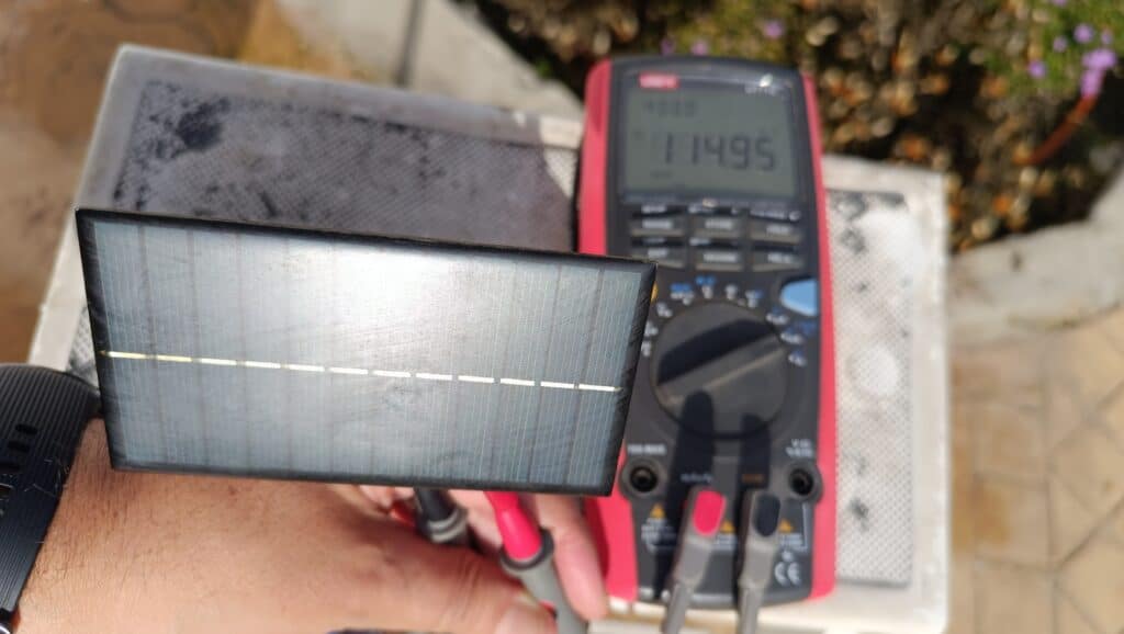 Célula solar rectangular sostenida por una mano con sondas conectadas y un multímetro digital rojo en segundo plano que marca aproximadamente 11,5 V, tomada en un entorno exterior.