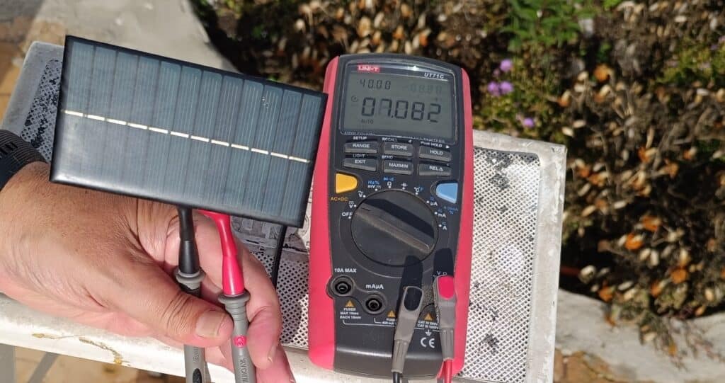 Mano sujetando un pequeño panel solar rectangular conectado con cables rojo y negro a las puntas de un multímetro digital rojo colocado sobre una superficie blanca, con vegetación de fondo y la pantalla del multímetro mostrando una lectura.